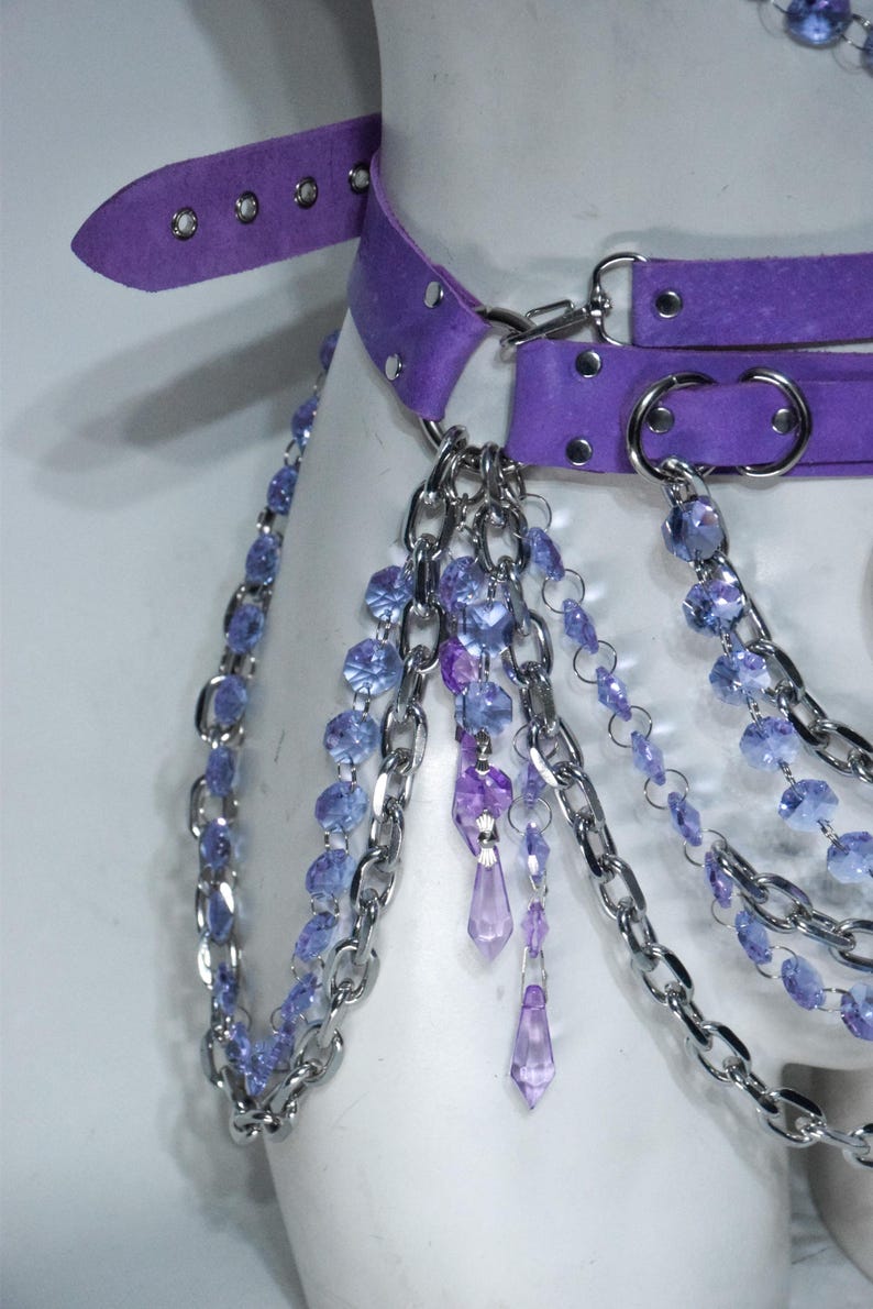 May include: A purple suede belt featuring silver-toned metal hardware, including circular rings and a buckle. The belt is adorned with multiple draped silver chains and strands of faceted light blue and purple crystal-like beads and pendants.