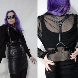 May include: A black leather harness with a large silver ring and straps. The harness is worn over a black mesh top and a black leather skirt.