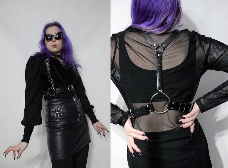May include: A black leather harness with a large silver ring and straps. The harness is worn over a black mesh top and a black leather skirt.