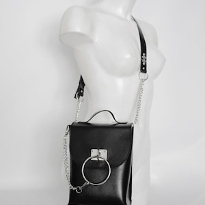 Puede incluir: Bolso bandolera de cuero negro con una correa de cadena plateada y un gran detalle de anillo plateado en la solapa delantera.