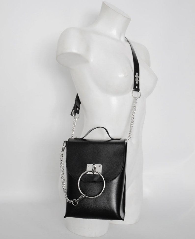 Puede incluir: Bolso bandolera de cuero negro con una correa de cadena plateada y un gran detalle de anillo plateado en la solapa delantera.