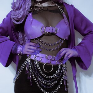 May include: A purple leather-like chest harness and waist belt set featuring silver-tone metal hardware, O-rings, and dangling purple crystal-style bead chains. The ensemble includes a black sheer top, a purple robe, and matching purple gloves.