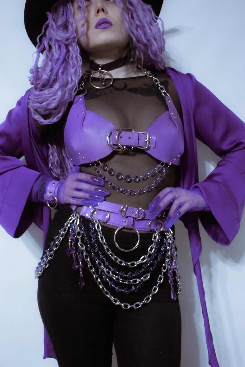 May include: A purple leather-like chest harness and waist belt set featuring silver-tone metal hardware, O-rings, and dangling purple crystal-style bead chains. The ensemble includes a black sheer top, a purple robe, and matching purple gloves.