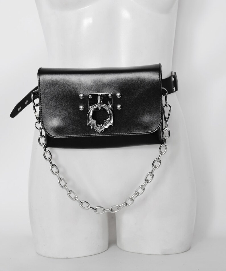 May include: Black leather fanny pack with a silver chain and a bat-shaped buckle. The bag has a flap with a silver ring.