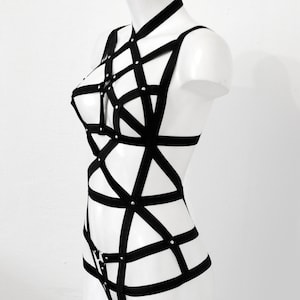 May include: Black body harness with a geometric design. The harness features multiple straps and silver-coloured rivets. The garment is displayed on a white mannequin against a white background. The harness is a lingerie item.