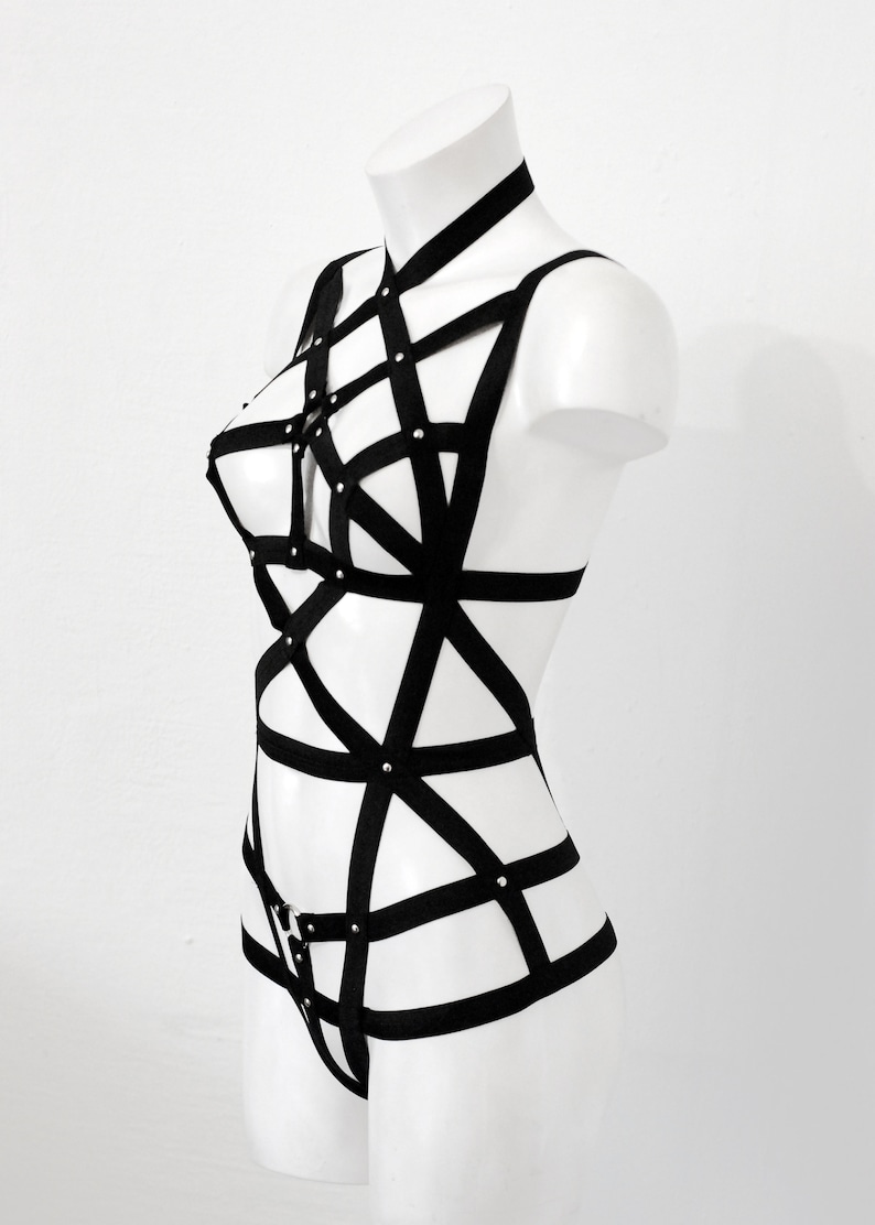 May include: Black body harness with a geometric design. The harness features multiple straps and silver-coloured rivets. The garment is displayed on a white mannequin against a white background. The harness is a lingerie item.