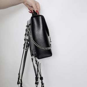 Puede incluir: Mochila de cuero negro con herrajes plateados y una correa de cadena. La mochila tiene m&uacute;ltiples correas y hebillas.