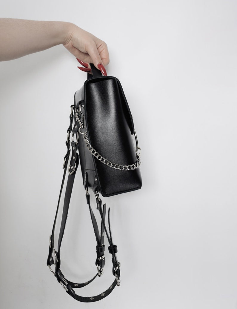 Puede incluir: Mochila de cuero negro con herrajes plateados y una correa de cadena. La mochila tiene m&uacute;ltiples correas y hebillas.
