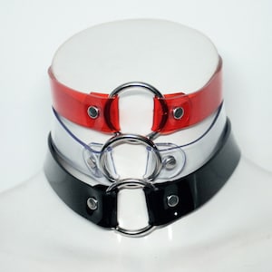 Puede incluir: Tres gargantillas con anillos plateados. La gargantilla superior es roja, la gargantilla del medio es transparente y la gargantilla inferior es negra.