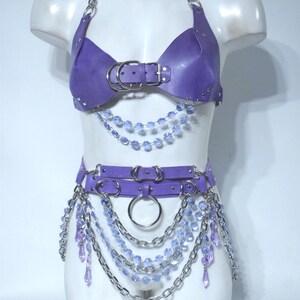 May include: A purple harness set featuring a halter-style top and a waist belt. The top has a central buckle and silver chain straps. The belt includes double straps with silver rings and chains, accented with dangling light purple crystal beads.