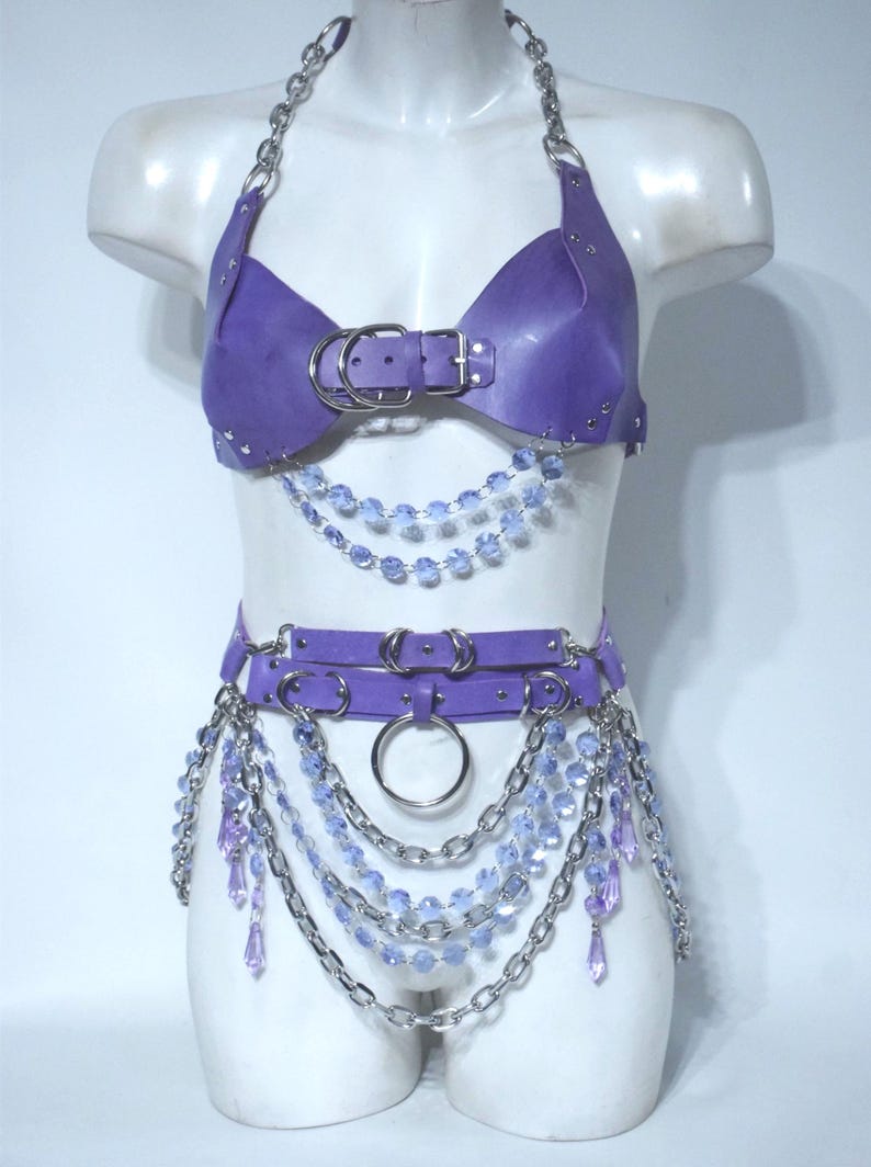 May include: A purple harness set featuring a halter-style top and a waist belt. The top has a central buckle and silver chain straps. The belt includes double straps with silver rings and chains, accented with dangling light purple crystal beads.