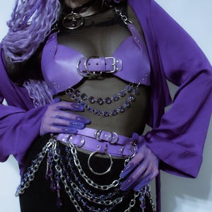 May include: A purple faux leather chest harness and matching waist belt featuring silver-tone metal buckles, O-rings, and layered chains with dangling purple crystal-like beads. The set is worn over a black sheer top and a black skirt with a purple jacket.