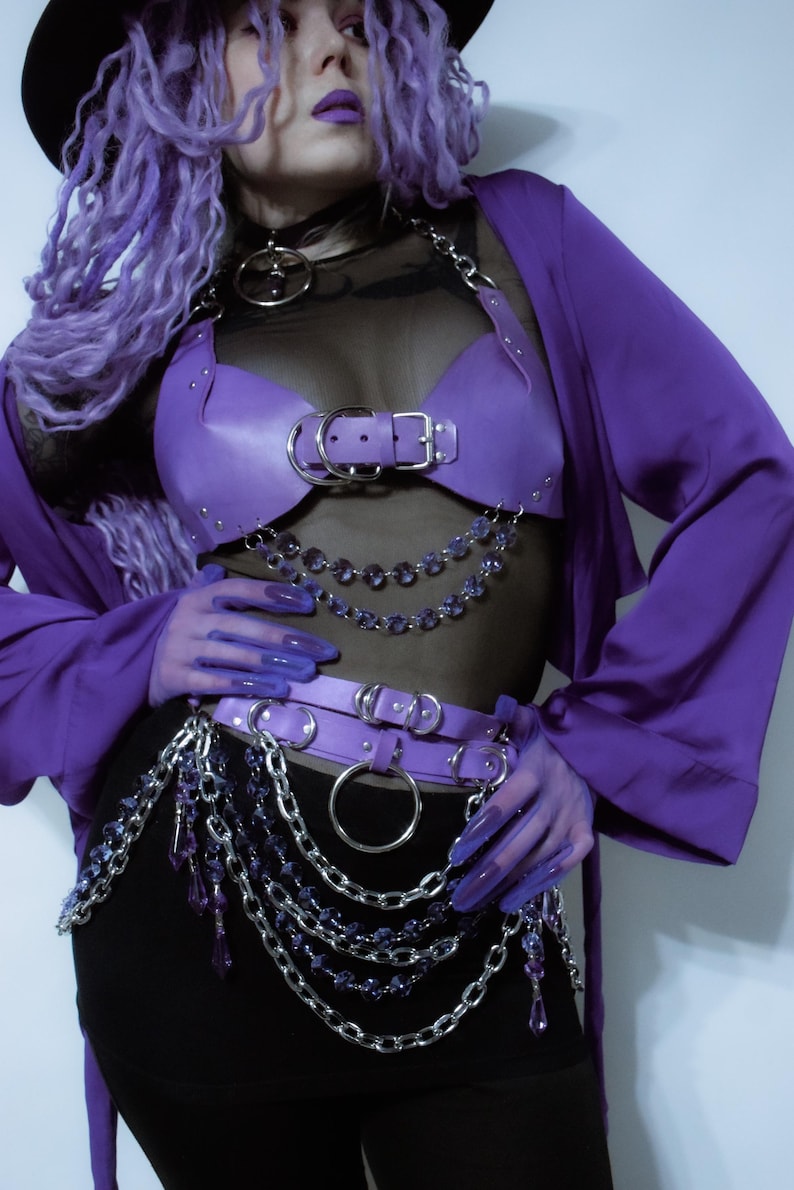 May include: A purple faux leather chest harness and matching waist belt featuring silver-tone metal buckles, O-rings, and layered chains with dangling purple crystal-like beads. The set is worn over a black sheer top and a black skirt with a purple jacket.