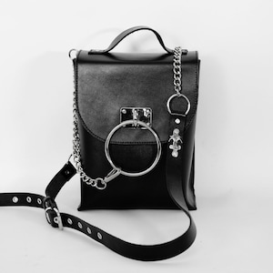 Puede incluir: Un bolso bandolera de cuero negro con una correa de cadena plateada y un gran detalle de anillo plateado. El bolso tiene un cierre de solapa y un peque&ntilde;o bolsillo en la parte delantera.