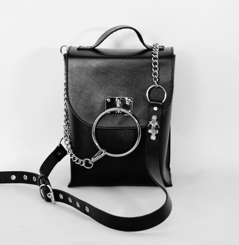 Puede incluir: Un bolso bandolera de cuero negro con una correa de cadena plateada y un gran detalle de anillo plateado. El bolso tiene un cierre de solapa y un peque&ntilde;o bolsillo en la parte delantera.