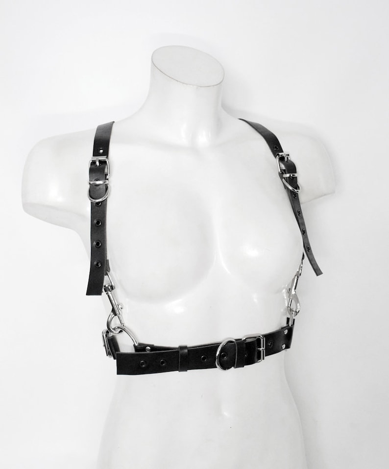 May include: A black leather harness with silver hardware. The harness has two straps that go over the shoulders and a belt that goes around the waist. The straps are adjustable and have buckles.