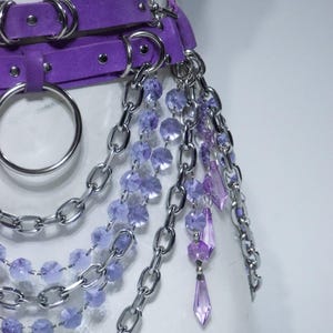 May include: A purple belt featuring silver-toned metal hardware, including large O-rings and multiple layered chains. The accessory is adorned with dangling strands of faceted light purple and dark purple beads, along with pointed crystal-like pendants.