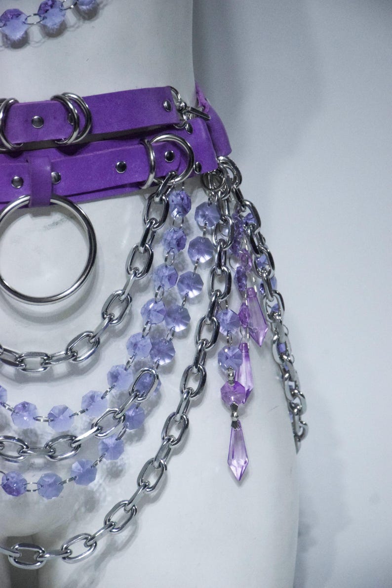 May include: A purple belt featuring silver-toned metal hardware, including large O-rings and multiple layered chains. The accessory is adorned with dangling strands of faceted light purple and dark purple beads, along with pointed crystal-like pendants.