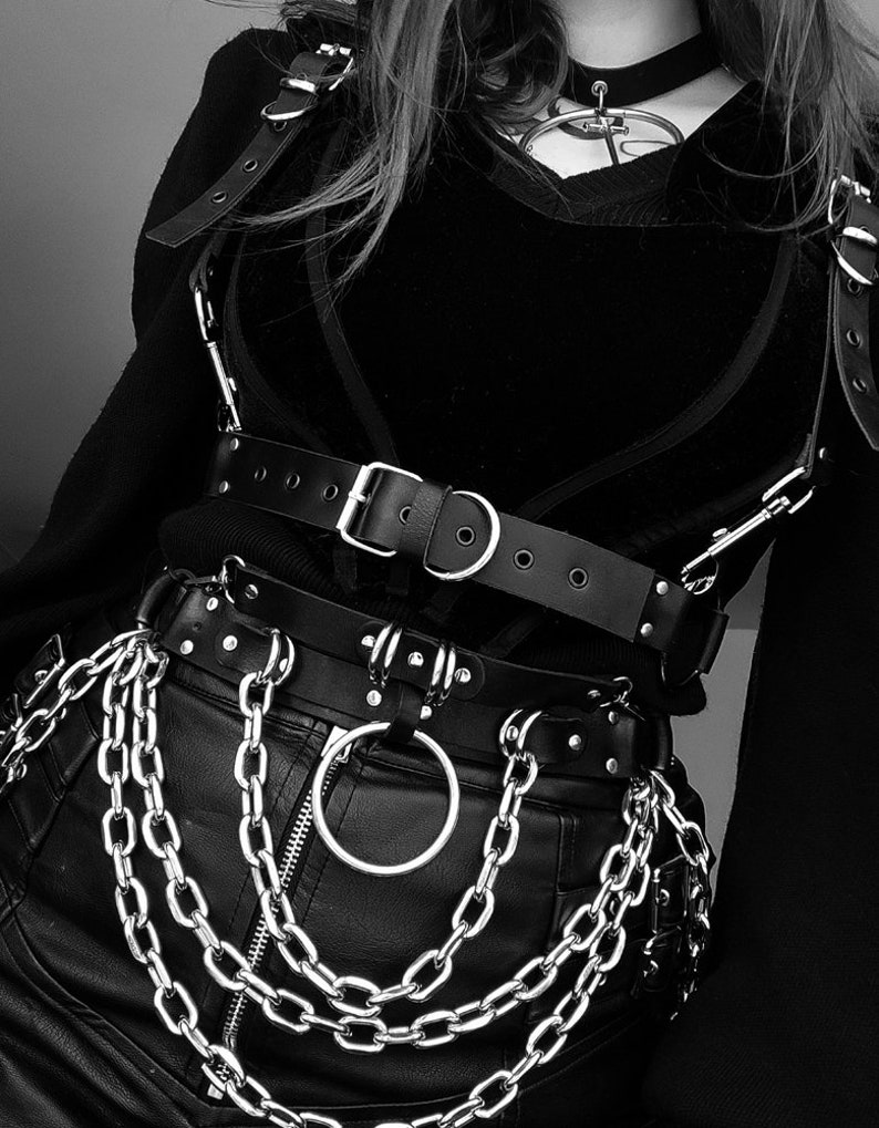 May include: A black leather harness with silver buckles and rings, worn over a black shirt. The harness is paired with a black leather belt with silver chains and a large silver ring.
