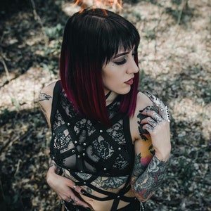 May include: A black lace top with a harness design, featuring silver studs and straps. The model has black hair with red highlights and multiple tattoos. The background is blurred with a forest setting.