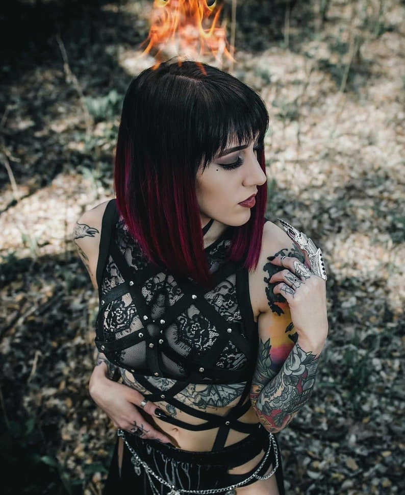 May include: A black lace top with a harness design, featuring silver studs and straps. The model has black hair with red highlights and multiple tattoos. The background is blurred with a forest setting.