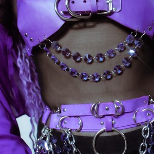 May include: A purple harness top and matching belt set featuring silver-toned metal buckles, rivets, and large circular rings. The garment is adorned with multiple draped chains of faceted purple crystal beads and thick silver-toned metal chain links.