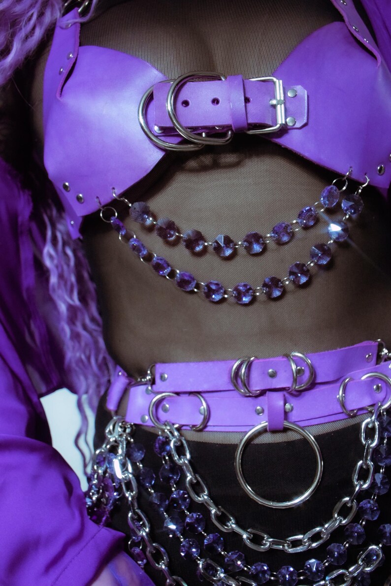 May include: A purple harness top and matching belt set featuring silver-toned metal buckles, rivets, and large circular rings. The garment is adorned with multiple draped chains of faceted purple crystal beads and thick silver-toned metal chain links.