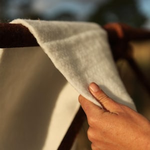 Puede incluir: Primer plano de una tela blanca y esponjosa que se sostiene con una mano. La tela está drapeada sobre una barra de metal oxidada.