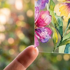 May include: A clear, rectangular piece of art featuring a watercolor floral design. The flowers are in shades of pink, purple, and yellow, with green leaves. The artwork is held up by a finger, with a blurred background of green foliage.