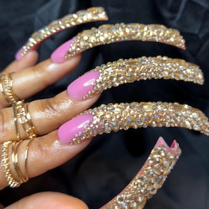 May include: Long, embellished fingernails with a pink base and gold-colored rhinestones. The nails are adorned with multiple gold rings. The overall aesthetic is glamorous and eye-catching, perfect for a special occasion or fashion statement.
