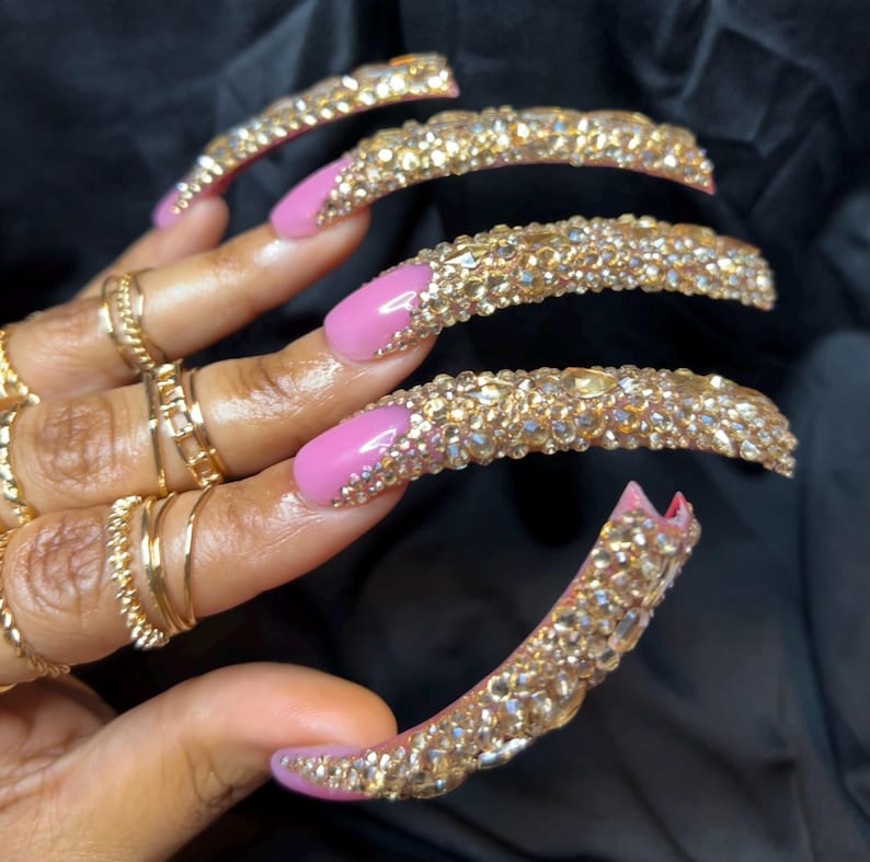 May include: Long, embellished fingernails with a pink base and gold-colored rhinestones. The nails are adorned with multiple gold rings. The overall aesthetic is glamorous and eye-catching, perfect for a special occasion or fashion statement.