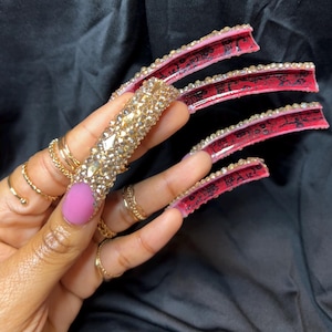 May include: A hand displaying long, embellished fingernails. One nail is covered in gold-colored rhinestones, while others feature red and black designs with clear rhinestones along the edges. The thumb nail is painted a solid pink color, and the hand is adorned with gold rings.