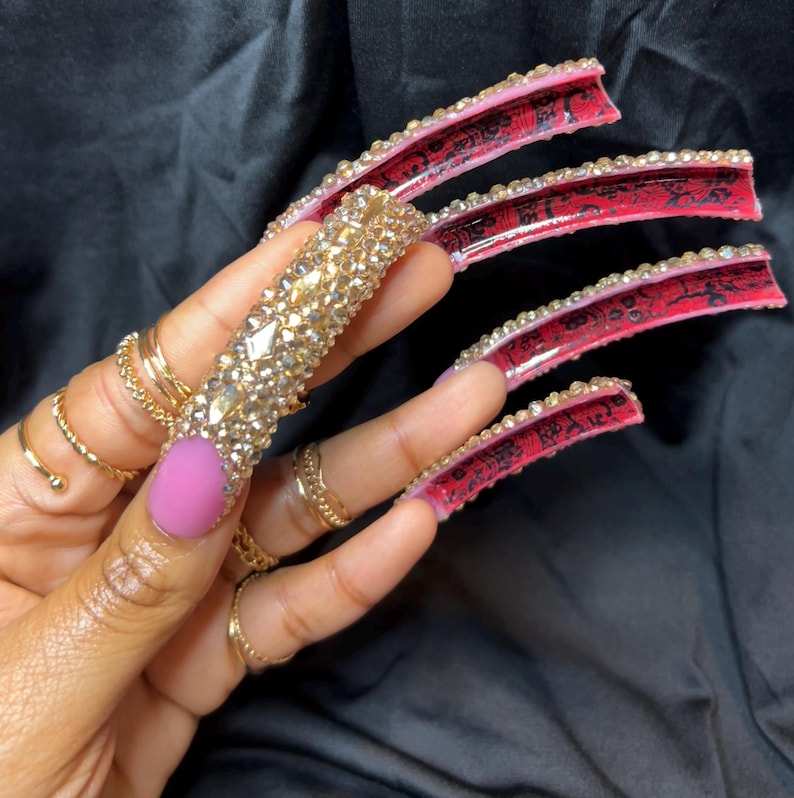 May include: A hand displaying long, embellished fingernails. One nail is covered in gold-colored rhinestones, while others feature red and black designs with clear rhinestones along the edges. The thumb nail is painted a solid pink color, and the hand is adorned with gold rings.