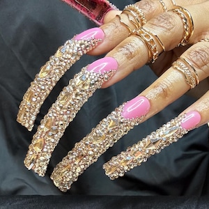 May include: Long, embellished artificial nails with a pink base and gold rhinestones. The nails are adorned with various sizes and shapes of sparkling crystals. Gold rings and bracelets complement the nail design.