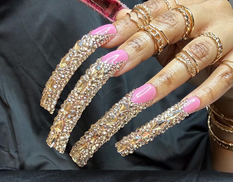 May include: Long, embellished artificial nails with a pink base and gold rhinestones. The nails are adorned with various sizes and shapes of sparkling crystals. Gold rings and bracelets complement the nail design.