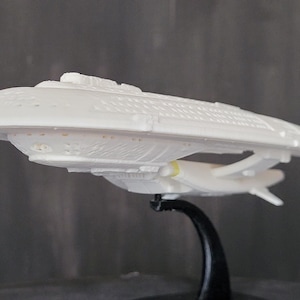 Peut inclure: Maquette blanche d'un vaisseau spatial, avec une coque principale en forme de soucoupe et deux nacelles secondaires. Le modèle est monté sur un support noir, avec des détails complexes.