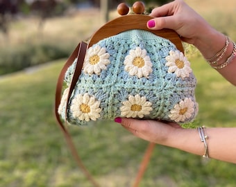 May include: A light green crocheted purse with a wooden frame and a brown strap. The purse is decorated with white crocheted flowers.