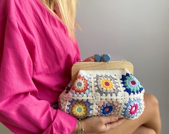 Puede incluir: Un bolso blanco de crochet con cuadrados de granny coloridos y un cierre de madera. El bolso lo lleva una persona que lleva una camisa rosa y pantalones cortos de mezclilla.
