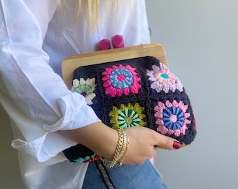 May include: A black crocheted clutch bag with a wooden frame and colorful floral accents. The bag has a clasp closure and is held by a person wearing a white shirt and denim shorts.