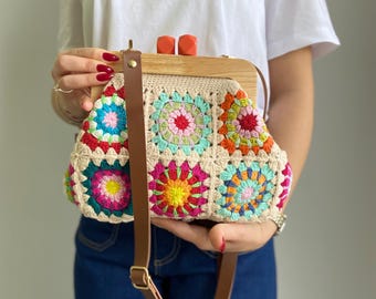 Puede incluir: Un bolso de crochet colorido con un marco de madera y una correa de cuero marrón. El bolso está decorado con un patrón floral y tiene un diseño único.