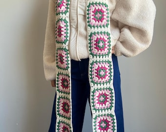 May include: A long, handmade crochet scarf featuring a repeating pattern of square motifs. Each square has a pink and green floral design, bordered by white and green. The scarf has a fringed edge and is displayed against a neutral background.