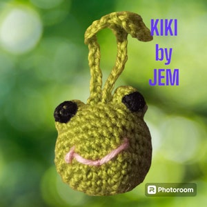 May include: A green crocheted frog with black eyes and a pink tongue. The frog is attached to a loop of green yarn. The text "KIKI by JEM" is visible in the background.