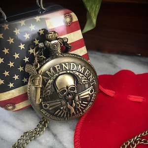 May include: A gold pocket watch with a skull and crossbones design. The watch face reads "Amendment II, Defending Liberty Since 1791".