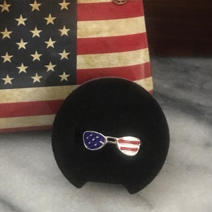 May include: A pair of silver sunglasses with an American flag design. The sunglasses are on a black velvet background.