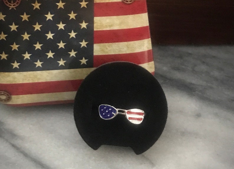 May include: A pair of silver sunglasses with an American flag design. The sunglasses are on a black velvet background.