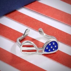 May include: A silver ring shaped like sunglasses with an American flag design. The sunglasses are red, white, and blue.