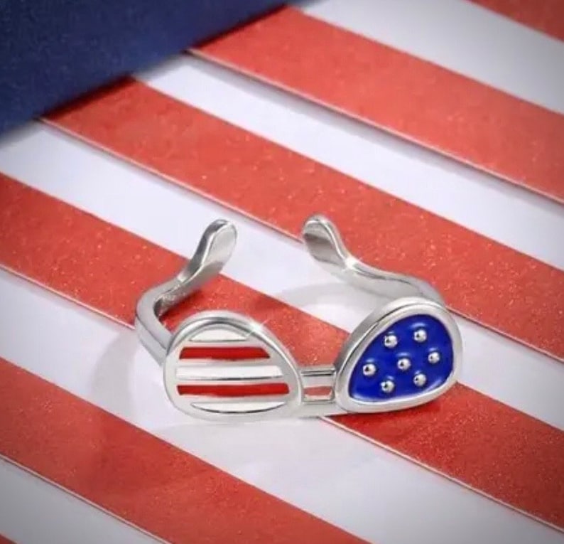 May include: A silver ring shaped like sunglasses with an American flag design. The sunglasses are red, white, and blue.