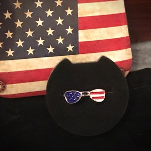 May include: A close-up of a small, silver pin shaped like a pair of sunglasses with red, white and blue enamel. The pin is on a black background.