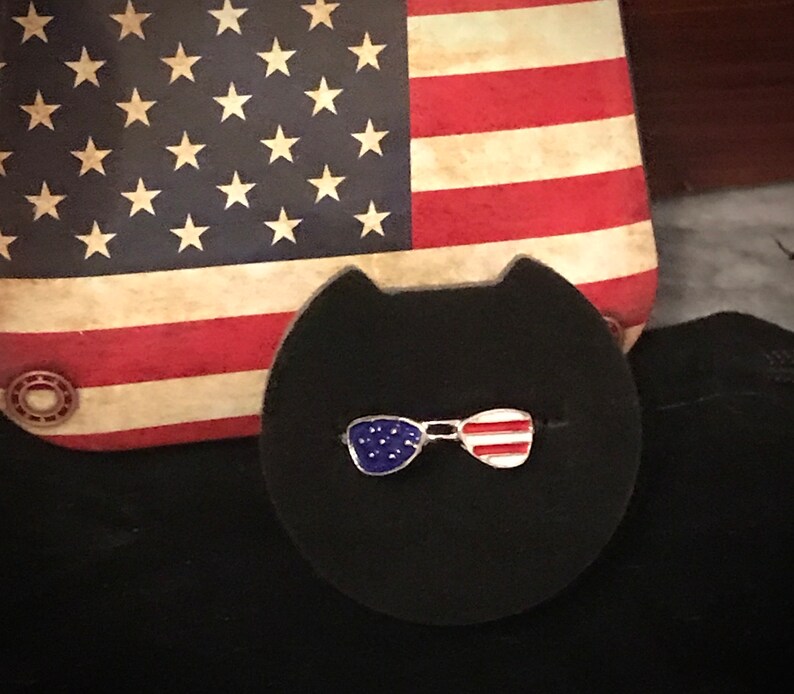May include: A close-up of a small, silver pin shaped like a pair of sunglasses with red, white and blue enamel. The pin is on a black background.