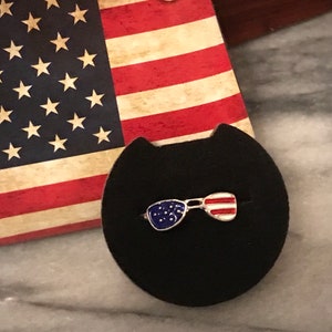 May include: A silver ring featuring an enamel design of sunglasses in blue and red. The sunglasses are decorated with stars and stripes, reminiscent of the Union Jack.
